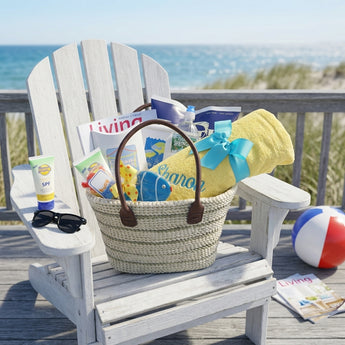 A Day at the Beach gift tote in woven rope with a yellow towel embroidered with a name, a magazine, water and snacks, on an Adirondack chair on a beach house deck with sunglasses and a beach ball