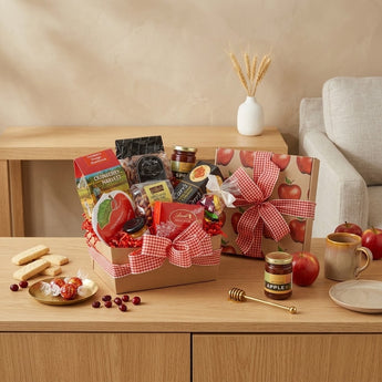 Autumn Harvest Gift Basket Regular size: a kraft gift box printed with red apples and tied with red gingham ribbon, filled with fall treats, with a matching closed apple print box beside it, styled on a light oak console table against a warm beige wall