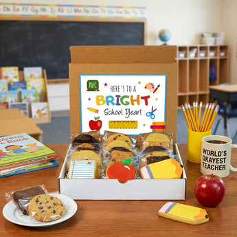 Back to school cookie and brownie box with a Here's to a Bright School Year card, wrapped chocolate chip cookies, fudge brownies and iced apple, pencil and notebook cookies, on a teacher's desk with pencils, picture books and a red apple