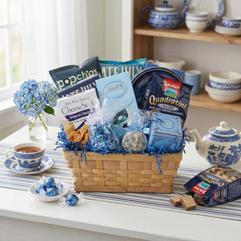 Blue Skies to You gift basket with blue crinkle shred and light blue bows, filled with Lindt Lindor, Loacker Quadratini, John Wm. Macy's CheeseSticks and popchips, on a cottage kitchen table with tea and blue hydrangeas