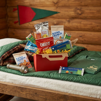 Red camp snack gable box with Cheez-It, Kayak Crunch snack mix and a Campfire Chocolate Bar, on a camp bunk with a green sleeping bag and a flashlight