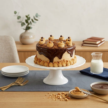 Chocolate peanut butter cake with ganache spiral, peanut butter rosettes and a crushed peanut border on a white pedestal stand, styled on an oak credenza with milk, peanut butter and gold forks