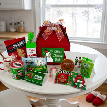 Personalized red Christmas gift box surrounded by holiday snacks and treats on a white kitchen table