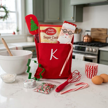 Christmas personalized baking kit with monogrammed red apron, cookie mix, spatula, whisk and cookie cutter on a white kitchen island