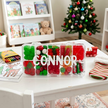 Personalized Christmas candy tackle box with a child's name on the lid and red and green candies on a playroom table by the tree
