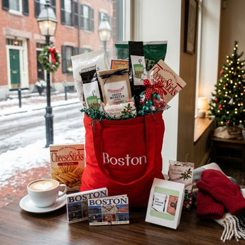 City Sidewalks Holiday Gift Tote Regular size: a red canvas tote printed with Boston in white script, filled with holiday snacks including CheeseSticks and a green coffee bag, with a Boston postcard book and a greeting card in front, styled on a dark wood table by a large window looking onto a snowy Beacon Hill brick street with a gas lamp