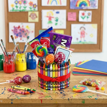 Crayon candy vase wrapped in crayons with a red ribbon, filled with Tootsie Pops, a rainbow swirl lollipop, Mike and Ike and jelly beans, on an art room table with paint jars and children's paintings