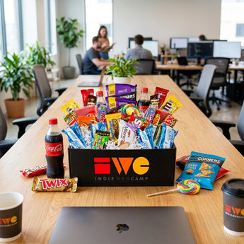 Deluxe snack attack box in a black branded corporate box overflowing with candy, Coca-Cola, Popcorners, Milano and Twix, on a communal table in a bright startup office