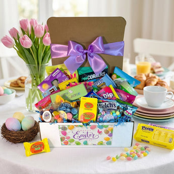 Easter snack box with a lavender bow and Happy Easter band, filled with Reese's eggs, Peeps, Welch's, Popcorners and pastel candy, on an Easter brunch table with pink tulips and dyed eggs