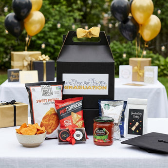 Graduation box of treats, a black gable box with a gold bow and Graduation label beside sweet potato chips, Popcorners, salsa, coffee and graduation cookies, on a backyard party gift table