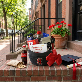 Hats-off graduation gift in a navy Boston baseball cap filled with snacks, a Coca-Cola and a Congrats Grad card with a red bow, on brownstone steps with peanuts and a grad cap