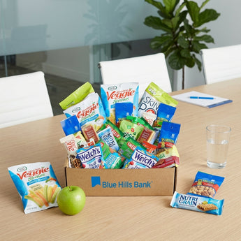 Healthy snack box in a kraft corporate box with a sample company logo, packed with Garden Veggie Straws, SkinnyPop, Planters, Welch's and Nutri-Grain, on a conference room table