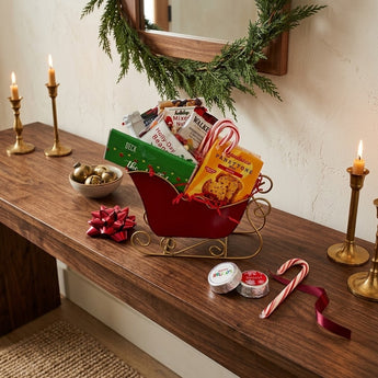 Holiday Cheer Sleigh gift basket in a red enamel sleigh with panettone, shortbread, mixed nuts and candy canes, styled on a walnut entryway console with brass candlesticks and a evergreen garland