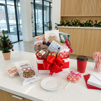 Holiday Crunch Gift Box Regular size: a white gift box tied with a big red ribbon bow, filled with holiday snacks including seasoned pretzels and CheeseSticks, styled on a white quartz office kitchen counter with tall windows onto a snowy city