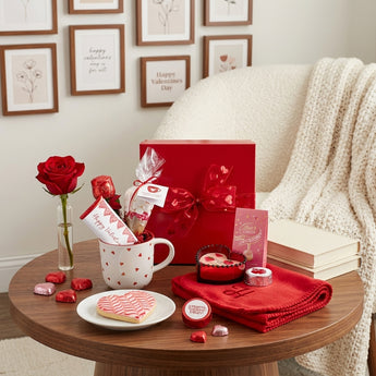 Hug in a Scarf Valentine's Day Gift Box, a red fleece scarf, heart mug with a chocolate rose, heart cookie and card, on a walnut side table in a cozy reading nook