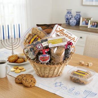 Kosher Nosh Gift Basket Regular size: a smaller natural willow basket with a clear tub of cookies, lavash crackers, a jar of preserves, molasses clove cookies and a bag of mixed nuts, styled on a light oak dining table with a white table runner embroidered with blue Hanukkah motifs