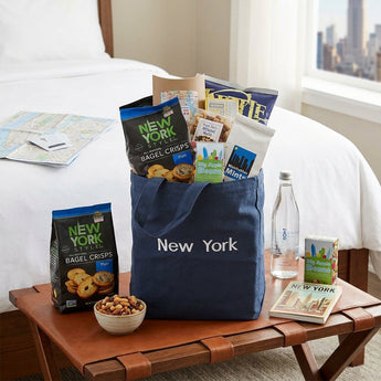 NY City Sidewalks Gift Basket, a navy New York tote filled with bagel crisps, Big Apple Beans, nuts and snacks, on a luggage rack in a SoHo hotel room