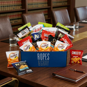 Office break snack box in a navy corporate box with a sample company name, packed with Goldfish, Cheez-It, Kettle chips and Garden Veggie Straws, on a walnut boardroom table