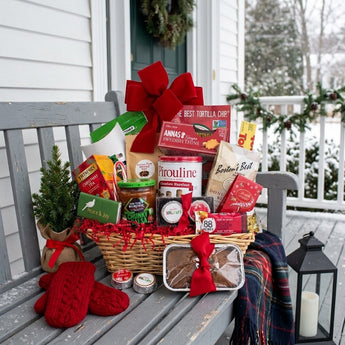 Peace and Joy holiday gift basket in a willow basket with a large red bow and snacks on a snowy front porch bench