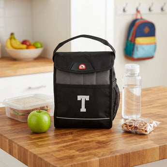 Black Igloo insulated lunch bag embroidered with a white letter T, on a butcher block kitchen counter with a sandwich, green apple, water bottle and pretzels