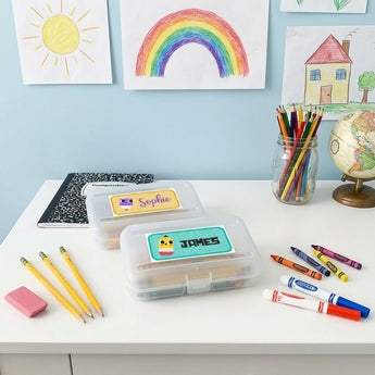 Two personalized clear pencil boxes with name labels reading Sophie and James, on a white kids homework desk with pencils, crayons, markers, an eraser and children's drawings