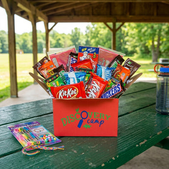 Snack attack box in a red camp branded box packed with Kit Kat, Snickers, Reese's and candy, on a green picnic table under a summer camp pavilion