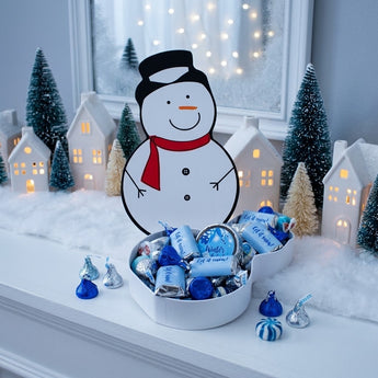 Snowman shaped candy box filled with blue and silver winter candy on a mantel with a snowy village