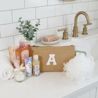 Stress relief spa package with a burlap cosmetic bag embroidered with the letter A, body wash, Honest lotion and body wash, a candle, a mesh pouf and tea, on a white bathroom vanity with a rolled towel and eucalyptus