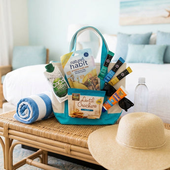 Summertime Amenity Gift, an aqua tote of trail mix, lentil crackers, coconut water, sunscreen and summer snacks, on a rattan bench in a coastal hotel room
