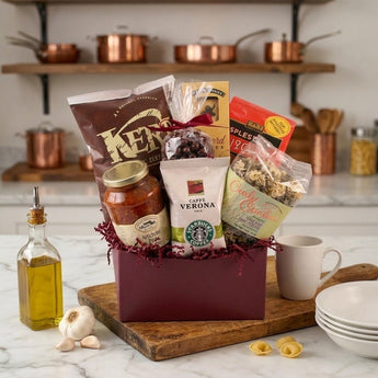 The North Ender Gift Basket Regular size: a burgundy gift box with red crinkle, filled with Italian treats including a STARBUCKS CAFFE VERONA coffee bag, a jar of pasta sauce, curly pasta and Kettle chips, styled on a white marble kitchen counter with copper pots on open wooden shelves