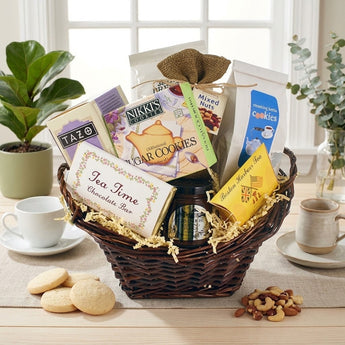 The Steaming Kettle Tea Gift Basket Regular size: a smaller dark brown wicker basket with natural crinkle, filled with TAZO tea, a Tea Time Chocolate Bar, Nikki's sugar cookies, Steaming Kettle cookies, Boston Harbor Tea and a bag of mixed nuts, styled on a light wood table by a white paned window with a linen runner