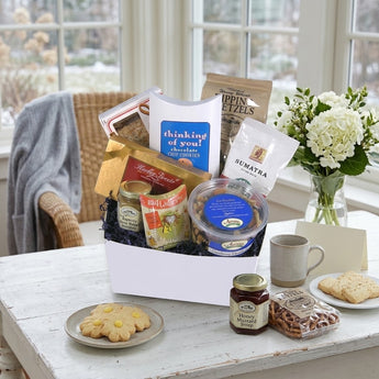 Thinking of You Gift Basket Regular size: a white gift box with navy crinkle, filled with comfort treats including Thinking of You chocolate chip cookies, Dipping Pretzels, Sumatra coffee, a jar of preserves and a tub of trail mix, styled on a whitewashed wooden table by a sunny window with white hydrangeas
