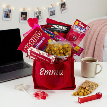Valentine's Day Personalized Gift Tote, a red tote embroidered with a name and filled with Haribo, Popcorners, candy and a heart lollipop, on a dorm room desk