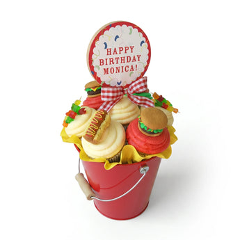 Decorative birthday bucket with cupcake-shaped cookies and a 'Happy Birthday Monica!' sign on a white background.