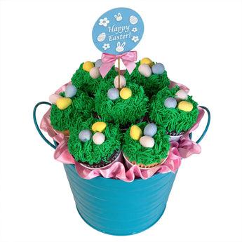 Easter-themed cupcake arrangement with green frosting, colorful eggs, and a 'Happy Easter!' sign in a blue bucket.