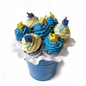 Cupcakes with blue and white frosting, topped with small toy dreidels, arranged in a blue bucket on a white background.