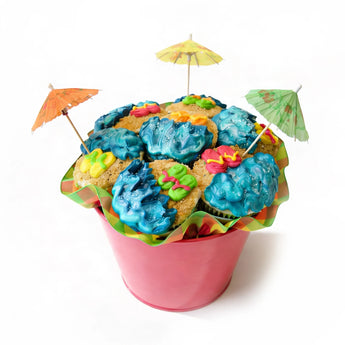 Colorful beach themed cupcakes with blue icing and decorative umbrellas in a pink bucket on a white background