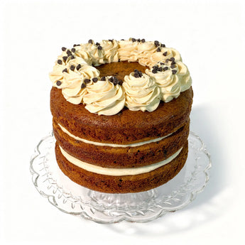 Three-layered cake with cream and chocolate chips on a clear stand against a white background