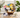 The Steaming Kettle Tea Gift Basket in a willow basket with a white teapot, teas, teapot sugar cookies and jam on a sunny table
