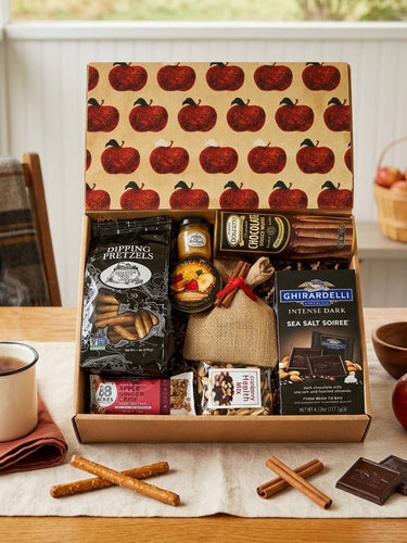 A Taste of Fall gift box with dipping pretzels, Ghirardelli dark chocolate, Dolcetto wafers, apple ginger crisp and cranberry health mix, styled on a farmhouse porch table with tea, apples and cinnamon