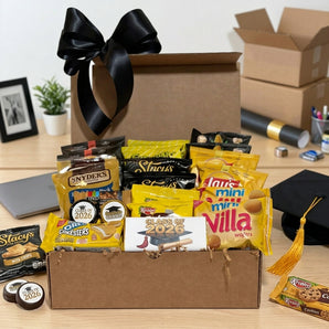 Graduation snack box with a black bow, packed with gold and yellow wrapped snacks, Class of 2026 chocolate bar and dipped Oreos, on a dorm desk among packing boxes with a grad cap
