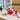 Holiday Crunch Gift Box Regular size: a white gift box tied with a big red ribbon bow, filled with holiday snacks including seasoned pretzels and CheeseSticks, styled on a white quartz office kitchen counter with tall windows onto a snowy city