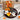 Eight hand iced Halloween sugar cookies on a dark plate, including two ghosts, a pumpkin, a black cat, a bat, candy corn, a spiderweb and a witch hat, in front of a kraft gift box with a Halloween pumpkin print sleeve and an orange satin bow on a bakery counter