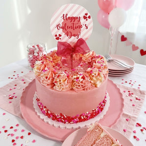 Valentine's Day sprinkles cake in soft pink buttercream with ruffled rosettes and a heart sprinkle band, styled on a party table with pink plates, candy and balloons