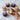 Hand holding a cupcake with purple flower frosting, surrounded by more cupcakes on a marble surface.